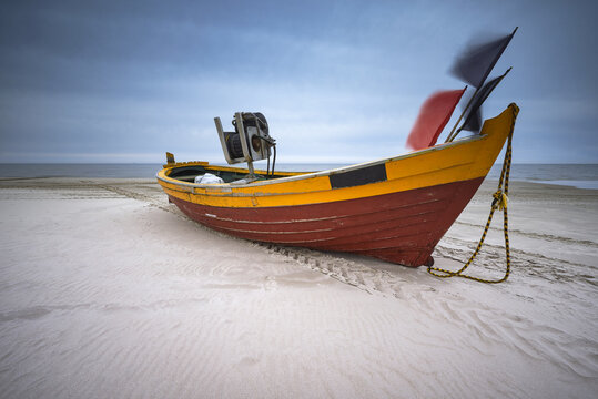 View of a colorful yellow and red wooden fishing boat resting on the sandy shore under a dramatic cloudy sky in Debki, Pomeranian Voivodeship, Poland.