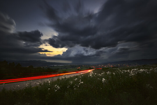 View of long exposure car light trails on a winding road near Czorsztyn under a dramatic stormy sky with dark clouds and distant town lights Czorsztyn, Lesser Poland, Poland.