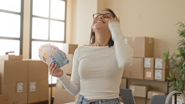 Woman holding a fanned stack of mexican pesos and parcels, hand raised in fist pump inside a building; small business success joy.