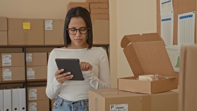 Woman tapping tablet while holding an open parcel box amid stacked shipping boxes and shelves in a packing building; focused productivity.