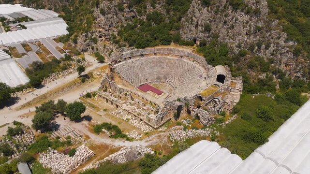 Demre, Turkey. Ancient Myra amphitheater, tiered stone seating carved into a mountainside, aerial view over the semi-circular cavea and surrounding Lycian rock formations under bright summer sun. Aer