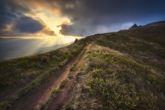 View of a winding dirt path on a grassy ridge of the Bieszczady Mountains under a dramatic sunset with sunbeams and dark clouds Bieszczady County, Podkarpackie Voivodeship, Poland.