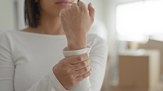 Young woman holding wrist and gripping sleeve beside stacked cardboard boxes inside building; moving stress.