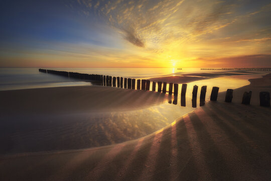 View of a golden sunset over a sandy beach with wooden groynes stretching into the calm sea in Chalupy, Pomeranian Voivodeship, Poland.