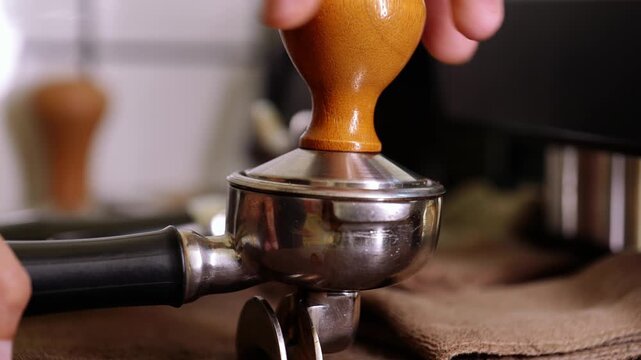 Close-up of a barista's hands pressing ground coffee with a wooden-handle tamper into a stainless steel portafilter, preparing espresso shot for a professional coffee machine.