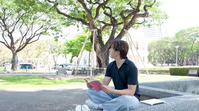 Thai Male Student Reading a Book in a Sunny University Campus Park