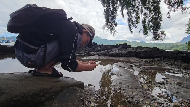 Young male traveler with a backpack crouching on a rocky shore to take a photograph of a water puddle reflecting the sky using his smartphone