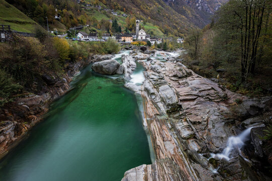View of Valle Verzasca with its emerald green river flowing over smooth rocks near a small village with a church tower in Verzasca, Ticino, Switzerland.