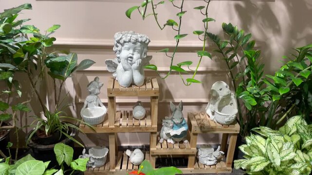 Small stone angel figurines on wooden tiered shelf surrounded by house plants