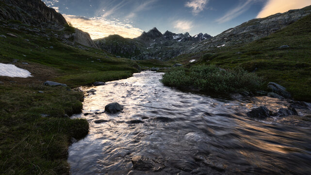 View of a mountain stream flowing through a rocky valley with green slopes and jagged peaks under a blue sky in the Aosta Valley, Italy.