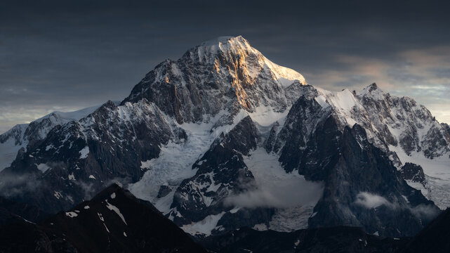 View of the majestic snow-covered peaks of the Mont Blanc massif illuminated by golden sunset light under a dramatic sky in Aosta Valley, Italy.