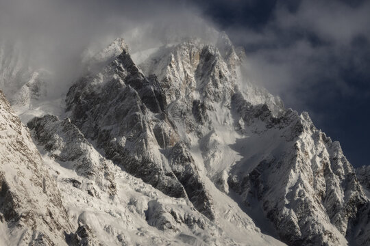 View of sharp snow-covered mountain peaks and rocky ridges partially hidden by thick clouds under a dark sky in Aosta Valley, Italy.
