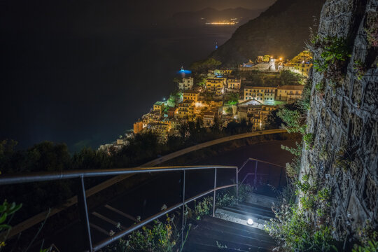 View of the illuminated village at night with stone stairs in the foreground and the dark sea in the background Riomaggiore, Liguria, Italy.