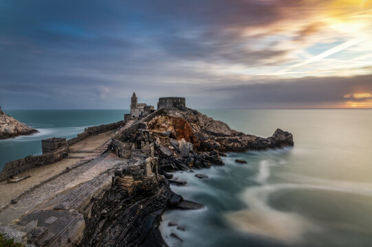 View of the Church of Saint Peter on a rocky promontory overlooking the sea with long exposure waves and sunset sky in Porto Venere, Liguria, Italy.
