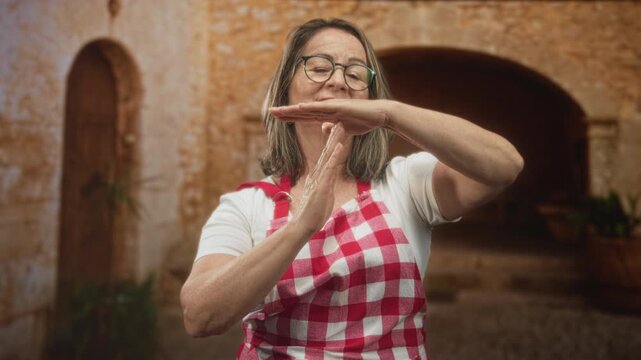 Hispanic woman cook forms time out gesture with hands wearing red checkered apron in building courtyard; home cooking confidence.