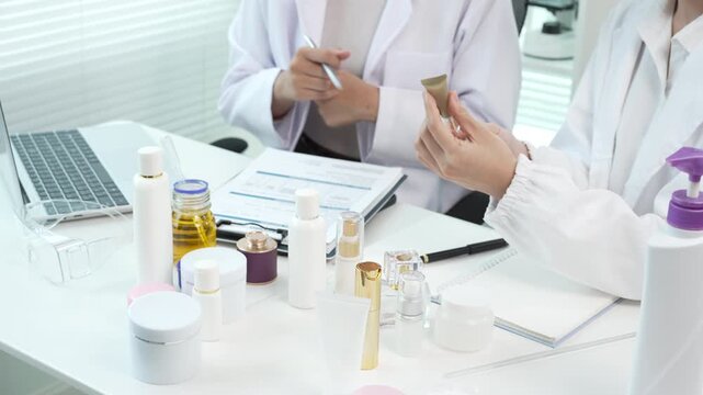 Cosmetic lab team reviewing skincare containers and product samples at a white desk with bottles, jars, notebook and laptop in a clean bright workspace.