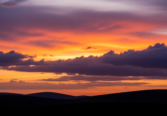 Fototapeta premium Vibrant sunset occurs over rolling hills with scattered clouds at dusk