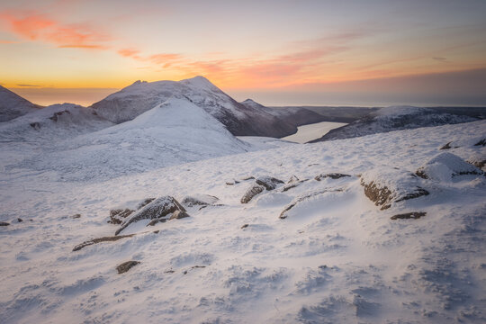 View of Slieve Meelmore and the snow-covered Mourne Mountains under a vibrant sunset sky with orange clouds and icy peaks in Newcastle, Northern Ireland, United Kingdom.
