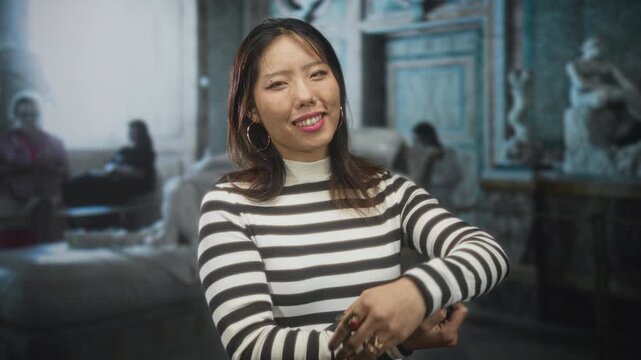 Woman holding tablet and touching ear while smiling in ornate building with sculptures and visitors, wearing striped turtleneck sweater and hoop earring; serenity curiosity.
