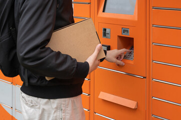 Man with backpack holding cardboard box. Entering PIN code on parcel locker keyboard. Receiving and sending parcels in an automated terminal © Gereish