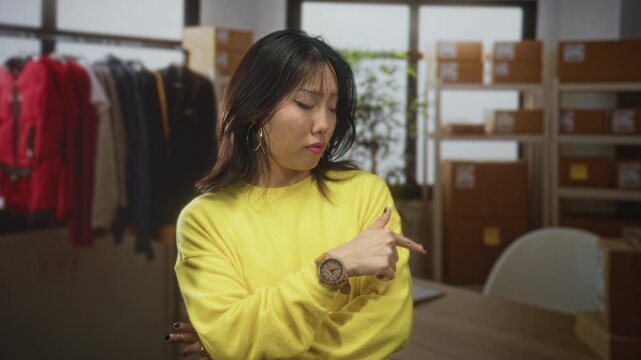 Young chinese woman points to wristwatch, checking time with finger gesture while wearing yellow sweater amid boxes and clothing racks in a packing building; thoughtful patience.