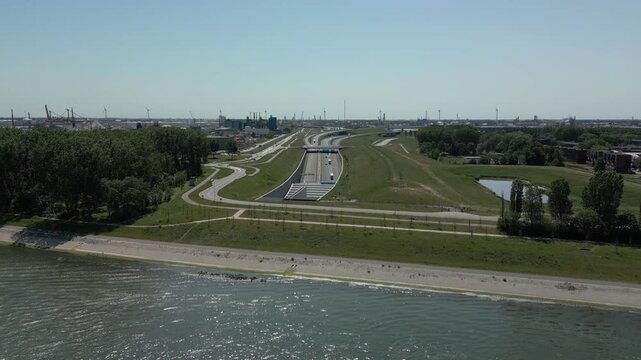 The Blankenburg Tunnel is embedded in a green waterfront zone, with the industrial silhouettes and wind turbines of the Port of Rotterdam in the background.