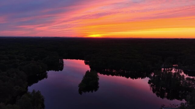 Drone view of colorful pink and orange sunset sky reflecting in dark lake water surrounded by forest in Harold Parker State Forest, Massachusetts