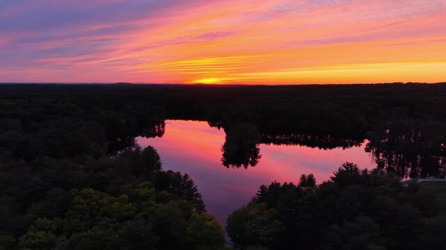 Drone view of colorful pink and orange sunset sky reflecting in calm lake surrounded by dark forest in Harold Parker State Forest, Massachusetts