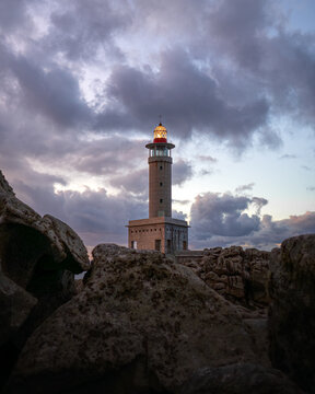 View of Faro de Punta Nariga lighthouse with its lantern lit against a dramatic cloudy sky and jagged coastal rocks in Malpica, A Coruna, Spain.