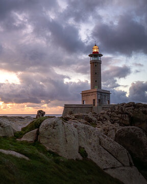 View of the Faro de Punta Nariga lighthouse standing on a rocky coastline under a dramatic sunset sky with glowing lantern light Malpica, A Coruna, Spain.