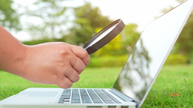Holding a magnifying glass to a laptop on green grass symbolizes attention to detail, tech curiosity, outdoor innovation, digital exploration, and critical thinking for smarter solutions.
