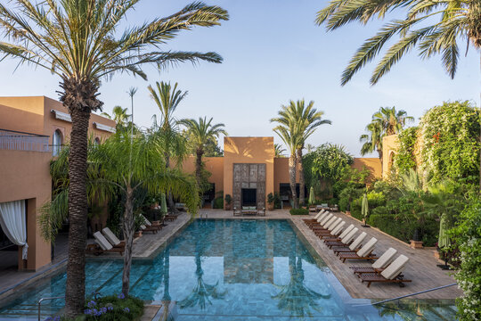 View of a luxury swimming pool surrounded by wooden lounge chairs, lush palm trees, and traditional terracotta architecture in Agadir, Souss-Massa, Morocco.