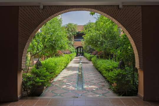 View of a lush Moroccan courtyard garden with a central water fountain and tiled paths framed by a brick archway Agadir, Souss-Massa, Morocco.