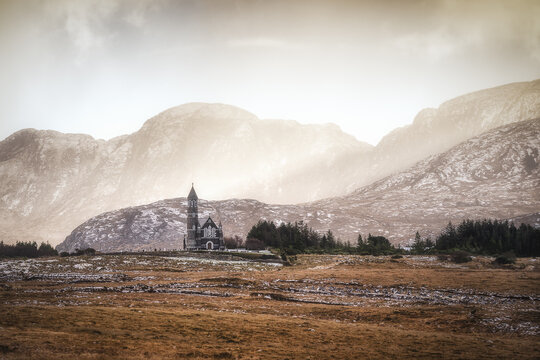 View of Sacred Heart Catholic Church at the foot of rugged mountains under dramatic misty light in County Donegal, Ireland.