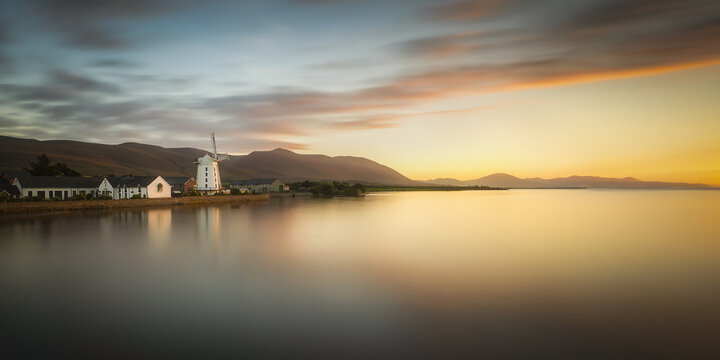 View of Blennerville Windmill and white houses along the calm waterfront with mountains under a sunset sky in Blennerville, County Kerry, Ireland.