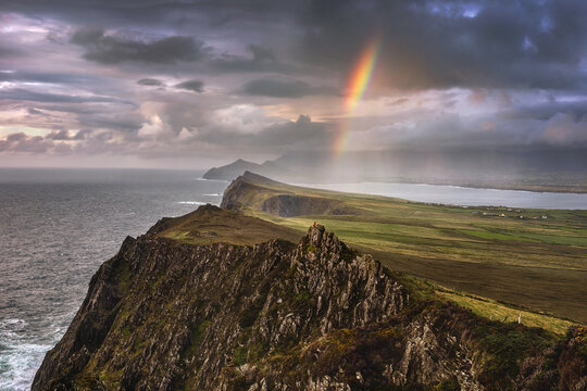 View of Sybil Head cliffs with a vibrant rainbow piercing through dramatic storm clouds over the rugged coastline and green fields in County Kerry, Ireland.