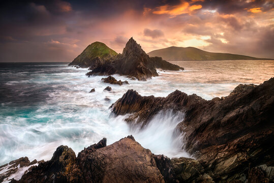 View of the jagged rocks and crashing waves at Dunmore Head under a dramatic sunset sky in County Kerry, Ireland.