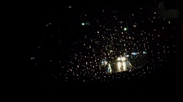 View of a car in the rear-view mirror from behind. View in the side rear view mirror. Nighttime road in the rain, headlights of oncoming cars, driving on the highway at night in the rain