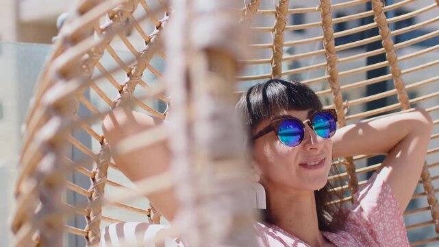 On a bright rooftop terrace, with city buildings in the background, a smiling woman wearing sunglasses sits comfortably in a round wicker hanging chair with white cushions
