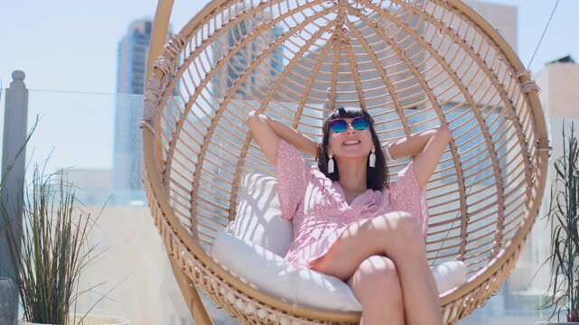 On a bright rooftop terrace, with city buildings in the background, a smiling woman wearing sunglasses sits comfortably in a round wicker hanging chair with white cushions