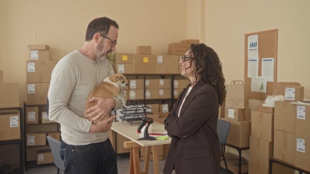 Man holding small dog and parcel while woman smiles amid stacked boxes in a packing building, suggesting daily delivery operations; partnership warmth.