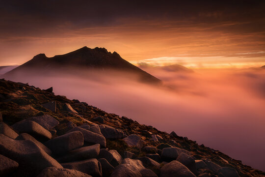 View of Slieve Bearnagh mountain peak rising above a thick layer of glowing pink mist and clouds during a vibrant orange sunset in Newry, Northern Ireland, United Kingdom.
