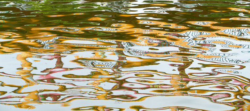 Close-up centered view of rippling water with highly distorted colorful reflections of trees and structures. Fragments of white, golden-yellow, blue, subtle pinks create fluid abstract patterns