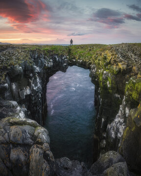 View of a person standing on the Gatklettur stone arch over the ocean under a dramatic sunset sky with pink clouds in Arnarstapi, Snaefellsbaer, Iceland.