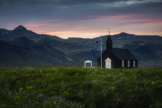View of Budakirkja black church standing in a field of wildflowers against a backdrop of rugged mountains and a colorful sunset sky in Budir, Snaefellsbaer, Iceland.
