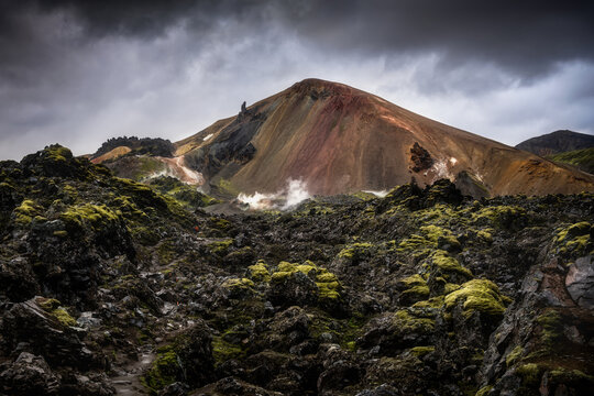 View of Landmannalaugar rhyolite mountains with moss-covered lava fields and geothermal steam under a dramatic cloudy sky Rangarthing ytra, Iceland.