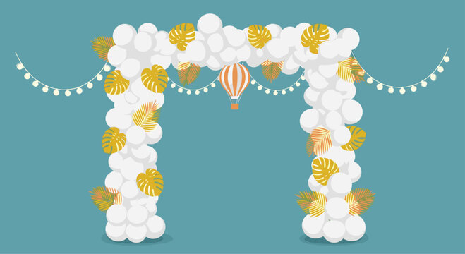 Festive decoration featuring a large arch made of white balloons adorned with golden tropical leaves and a small orange hot air balloon in the center.