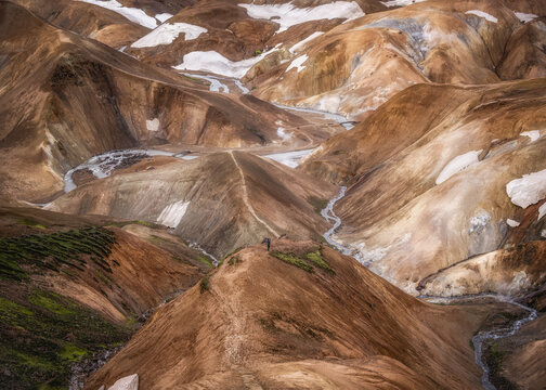 View of the rhyolite mountains of Kerlingarfjoll with snow patches, geothermal steam, and a person standing on a ridge, Hrunamannahreppur, Iceland.