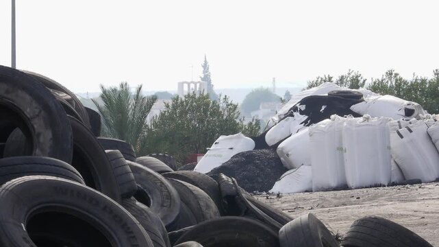 Static shot of a pile of recycled black rubber powder and large white industrial big bags stored outdoors at a tire recycling facility. Waste management and circular economy concept.