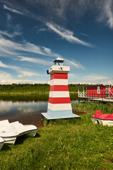 Obraz premium A red and white striped wooden lighthouse stands on grassy lake shore next to small pedal boats and a red boathouse. The scene shows calm reflective water, a
